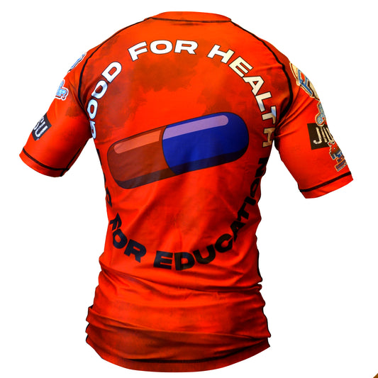 A person wearing the Retro Grappler Akira Grappler Rash Guard, a bright red polyester-spandex shirt featuring a large blue and red capsule graphic and “GOOD FOR HEALTH, GOOD FOR EDUCATION” printed in bold white and black letters on the back.