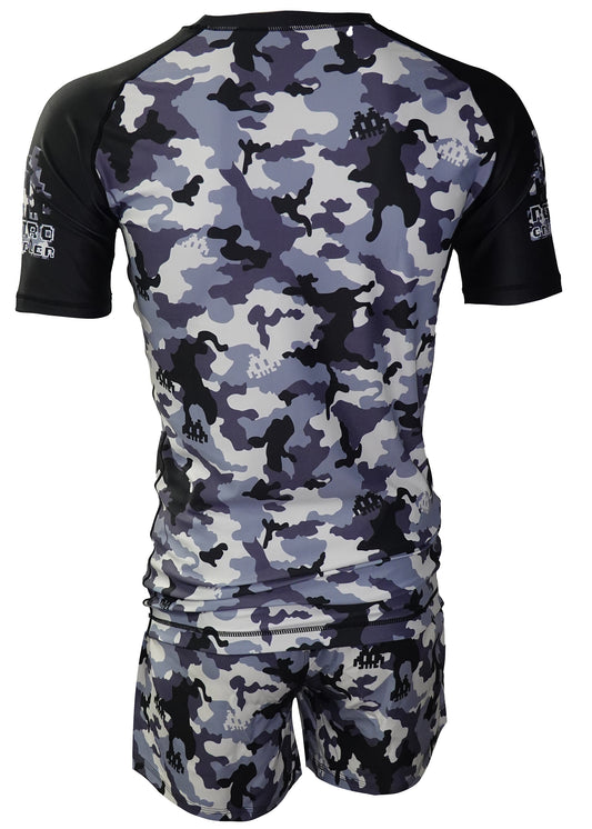 A person seen from the back wears the Retro Grappler Dark Woodland Camo Kit—a rash guard and matching shorts with a camo pattern featuring dinosaur silhouettes in gray, black, and white.