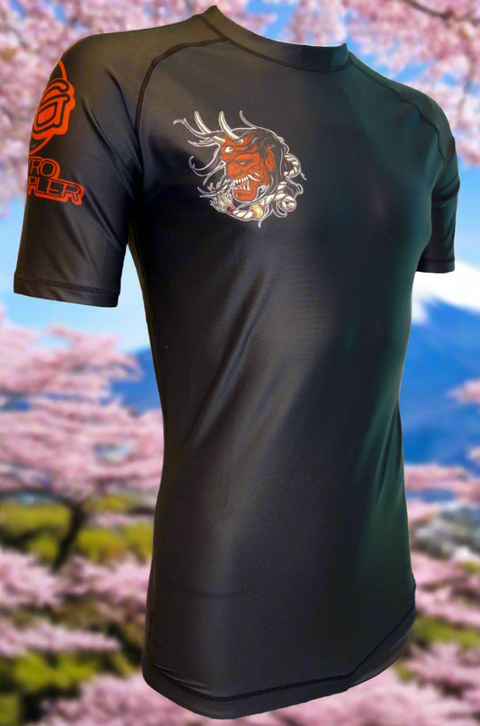 The Retro Grappler “Samurai Prodigy” Rash Guard is a black athletic shirt with Japanese Oni-inspired art on the chest and red logo text on the sleeve—ideal for jiu jitsu, shown against cherry blossoms and a blue sky.