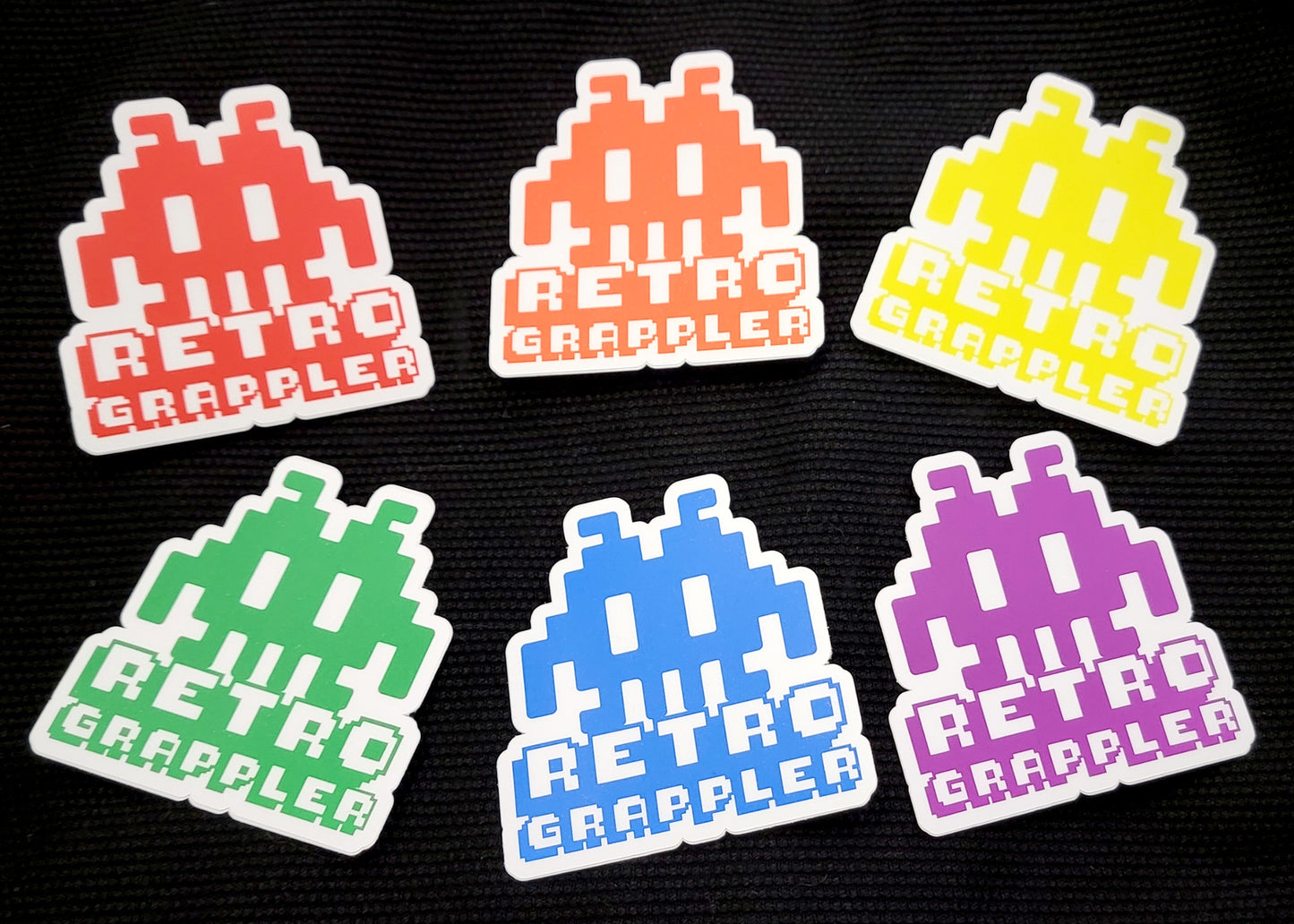 The Retro Grappler Sticker Pack by Retro Grappler includes six vinyl stickers with pixelated orange, yellow, green, blue, red, and purple alien designs and Retro Grappler text on a black textured background.
