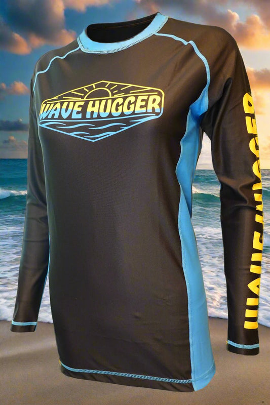 The Wave Hugger Blue Panel Rash Guard features long sleeves, fast-drying lycra, and “WAVE HUGGER” in yellow and blue on the chest and sleeve, shown against a sunset ocean beach background.
