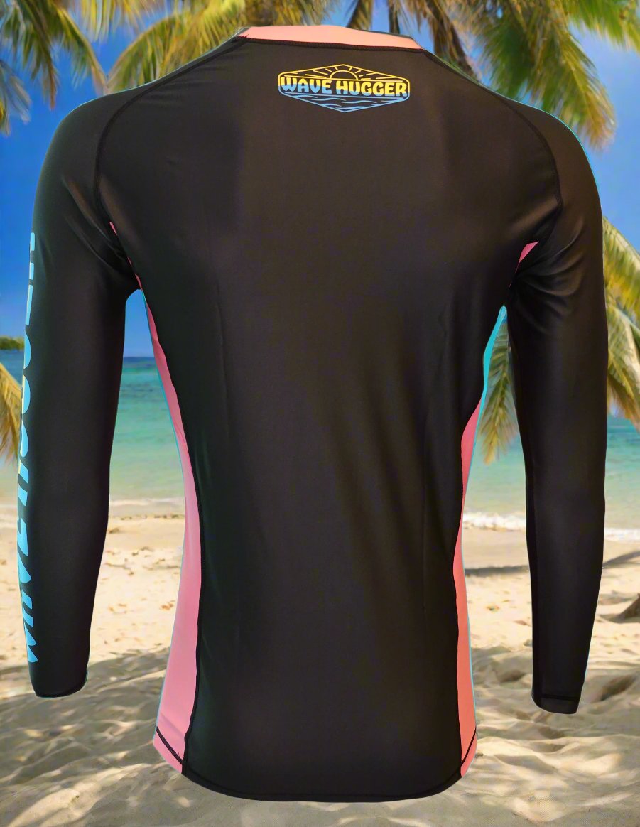 The Wave Hugger Pink Panel Rash Guard is a black and pink unisex long-sleeve, fast-drying lycra top with the Wave Hugger logo on the upper back, shown against a tropical palm beach backdrop.