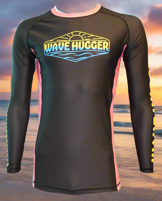 The Wave Hugger Pink Panel Rash Guard is a black, long-sleeve unisex lycra top with pink side panels and a yellow-blue Wave Hugger logo on the chest, shown against a beach sunset background.