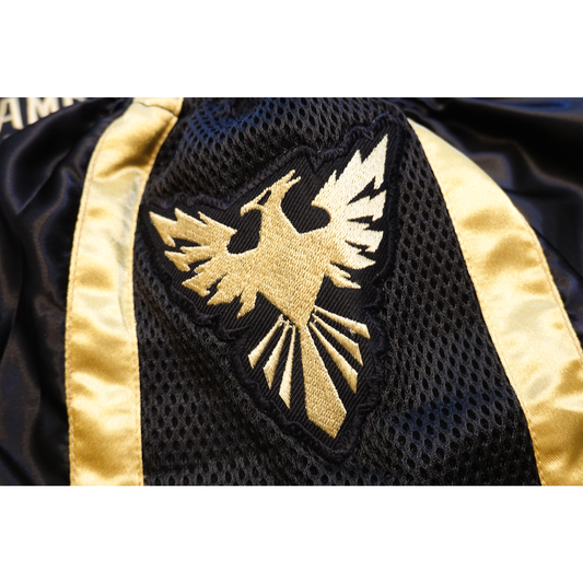 A close-up of a gold-bordered black fabric patch with a gold and white phoenix logo, sewn onto Combat Night Black Gold Premium Muay Thai shorts by Combat Night.